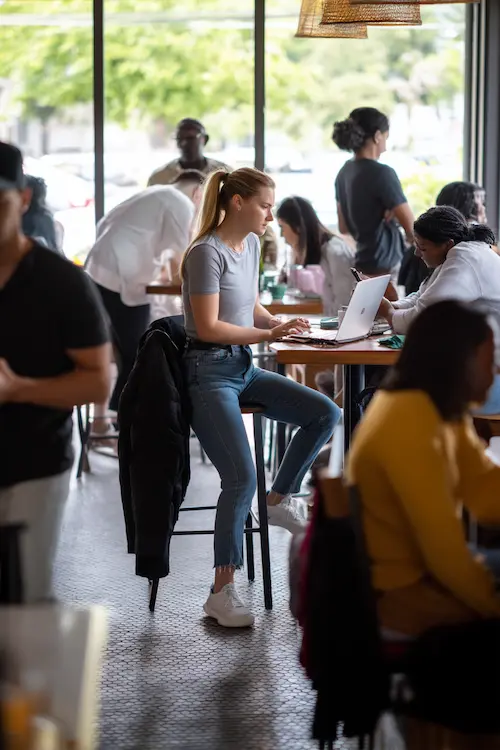 Een jonge vrouw in casual kleding werkt geconcentreerd op haar laptop in een levendig café, omringd door andere gasten die praten en werken.