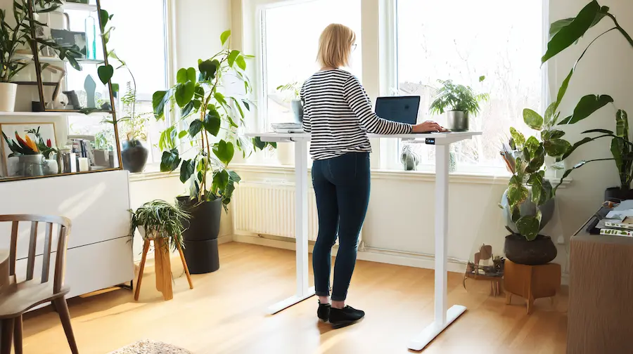 Thuiswerkplek met een elektrisch verstelbaar bureau, grote monitor en houten blad, aangevuld met een minimalistische kruk.