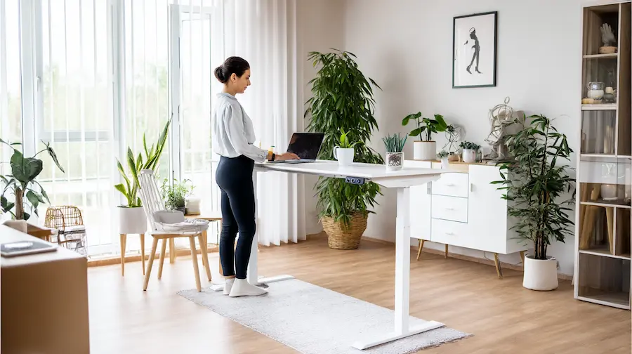 Vrouw werkt staand aan een elektrisch verstelbaar bureau in een modern ingerichte woonkamer met veel planten, natuurlijk licht en een laptop.