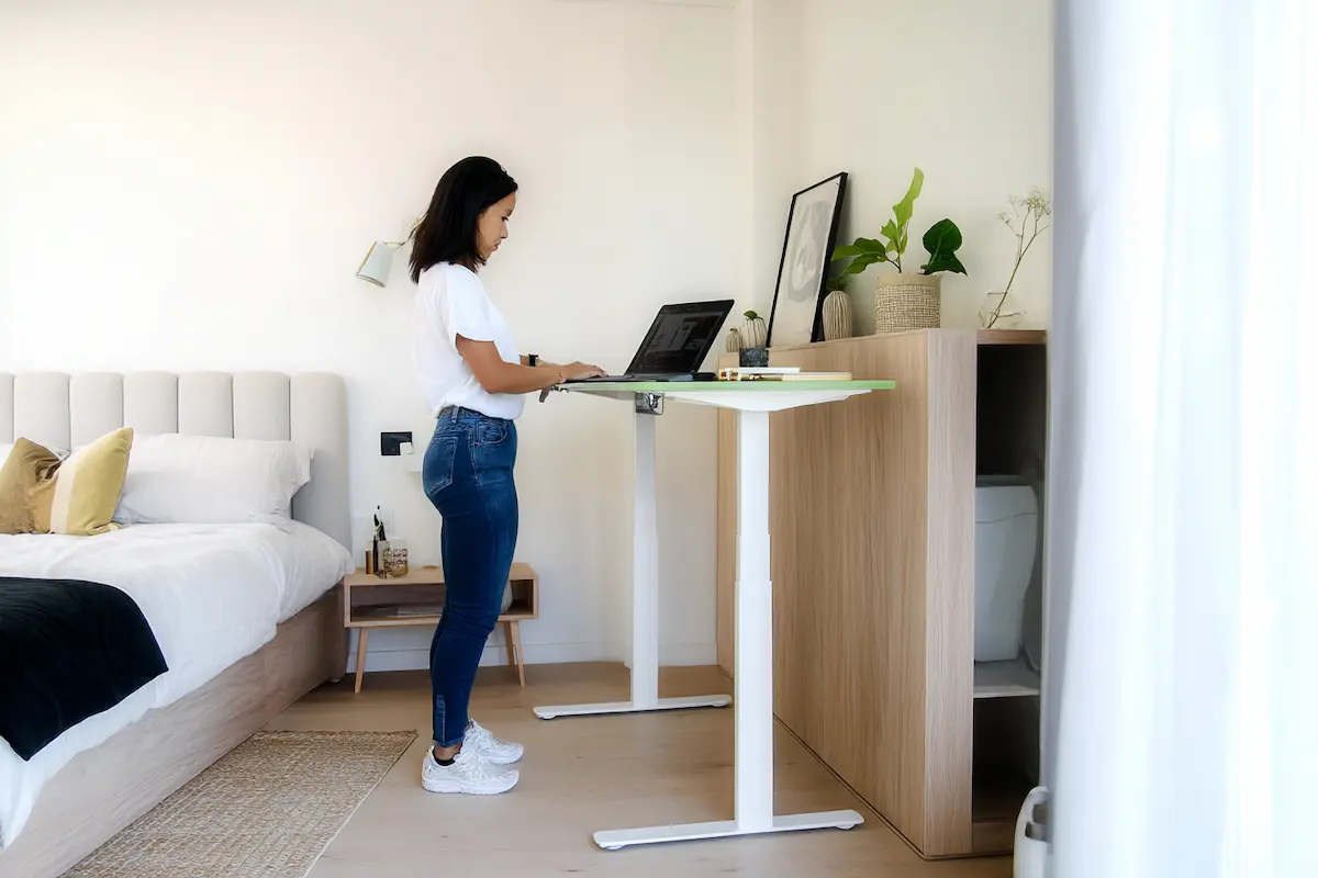 Vrouw werkt staand aan een elektrisch verstelbaar bureau bij het raam, met uitzicht op de buitenwereld en omgeven door planten.