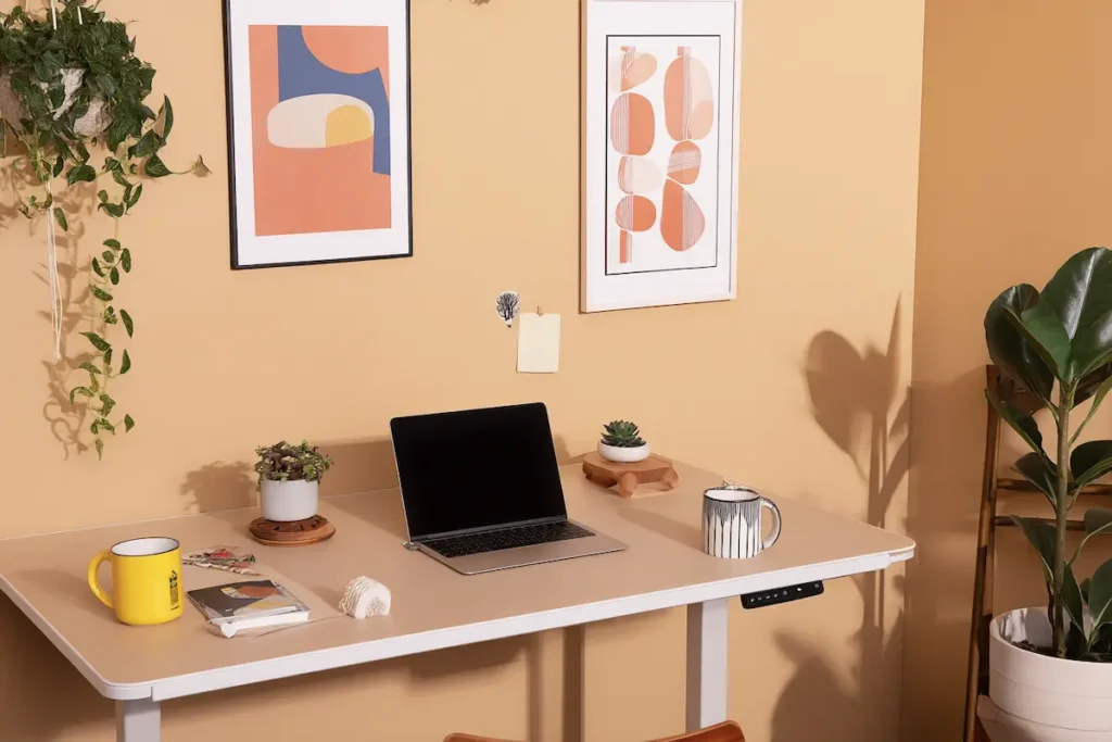 Elektrisch zit-sta bureau in een creatieve ruimte met beige muur, laptop, planten, koffiemokken en kleurrijke kunst aan de wand.