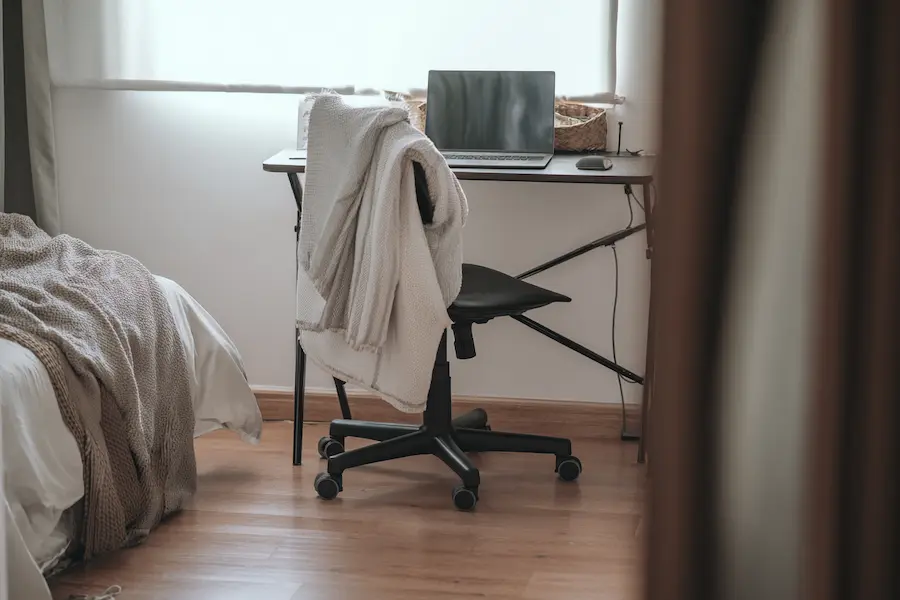 Een zwarte bureaustoel met een witte deken eroverheen gedrapeerd, aan een smal bureau met een laptop, naast een netjes opgemaakt bed met taupe en wit beddengoed.